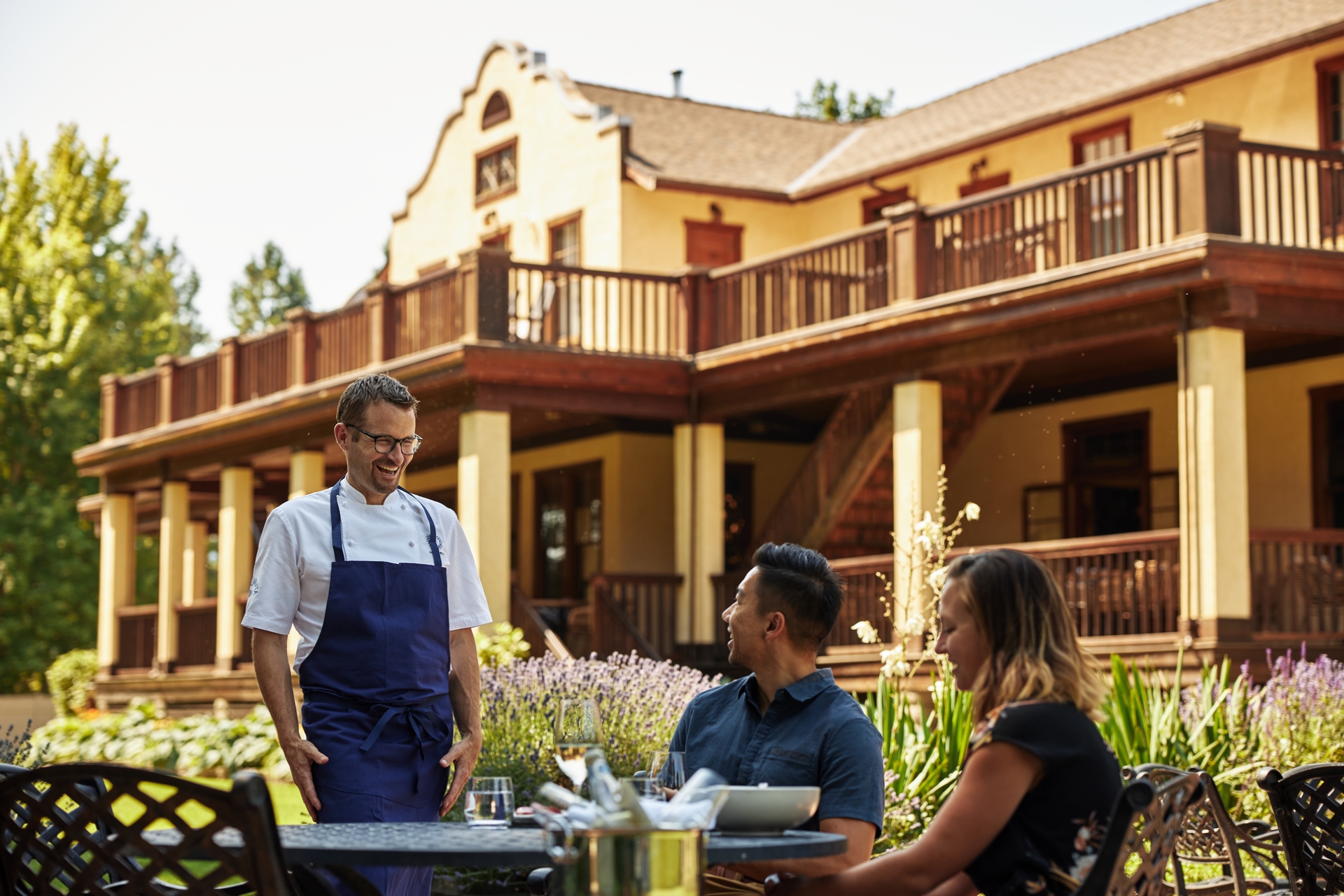 Chef Ned Bell describes the meal to a couple dining on the patio at the Naramata Inn. The building is two stories with a wrap around patio and gardens on the ground level.