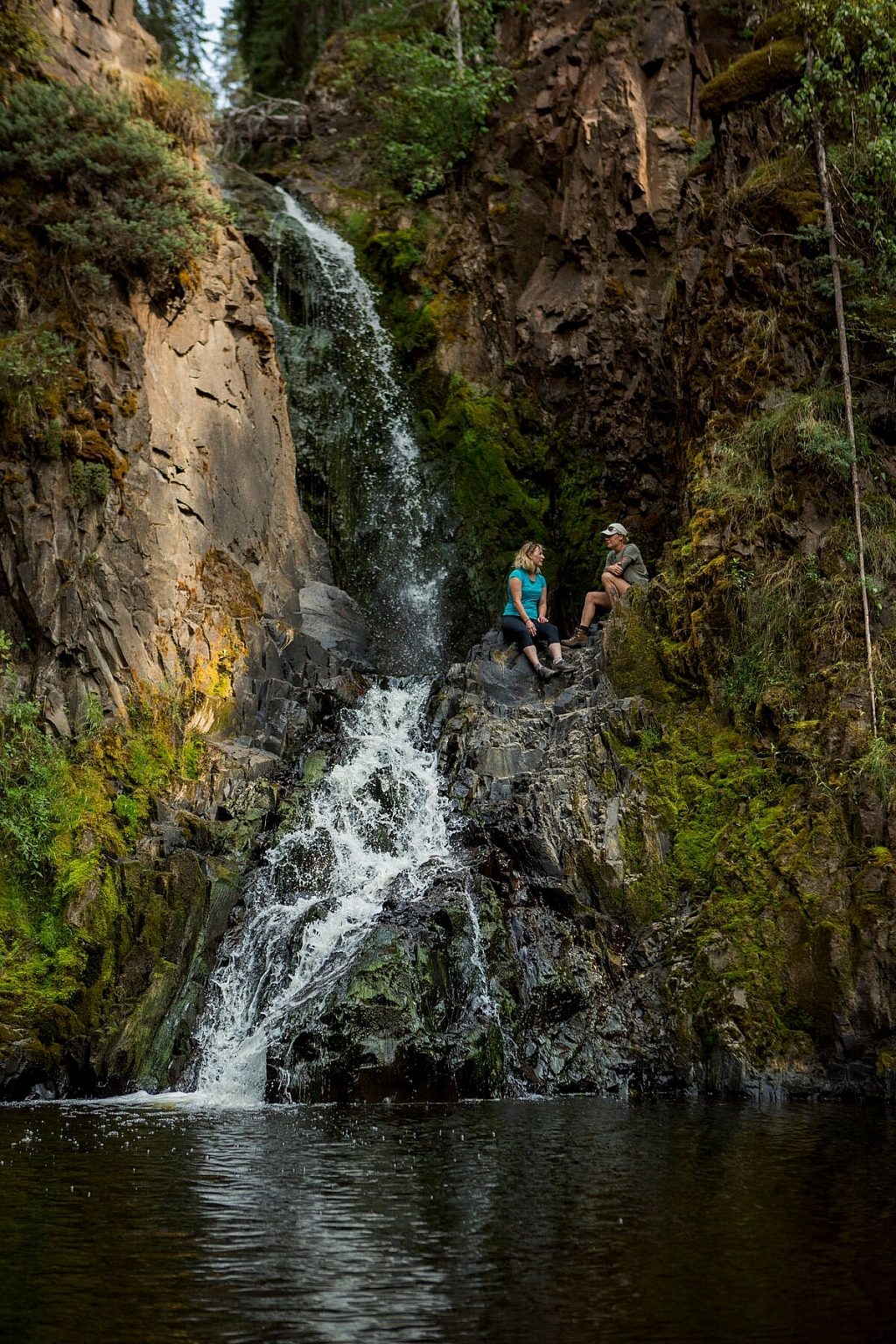 Two people sit beside Aitken Falls - a small waterfall trickling down the rocks into the lake below.