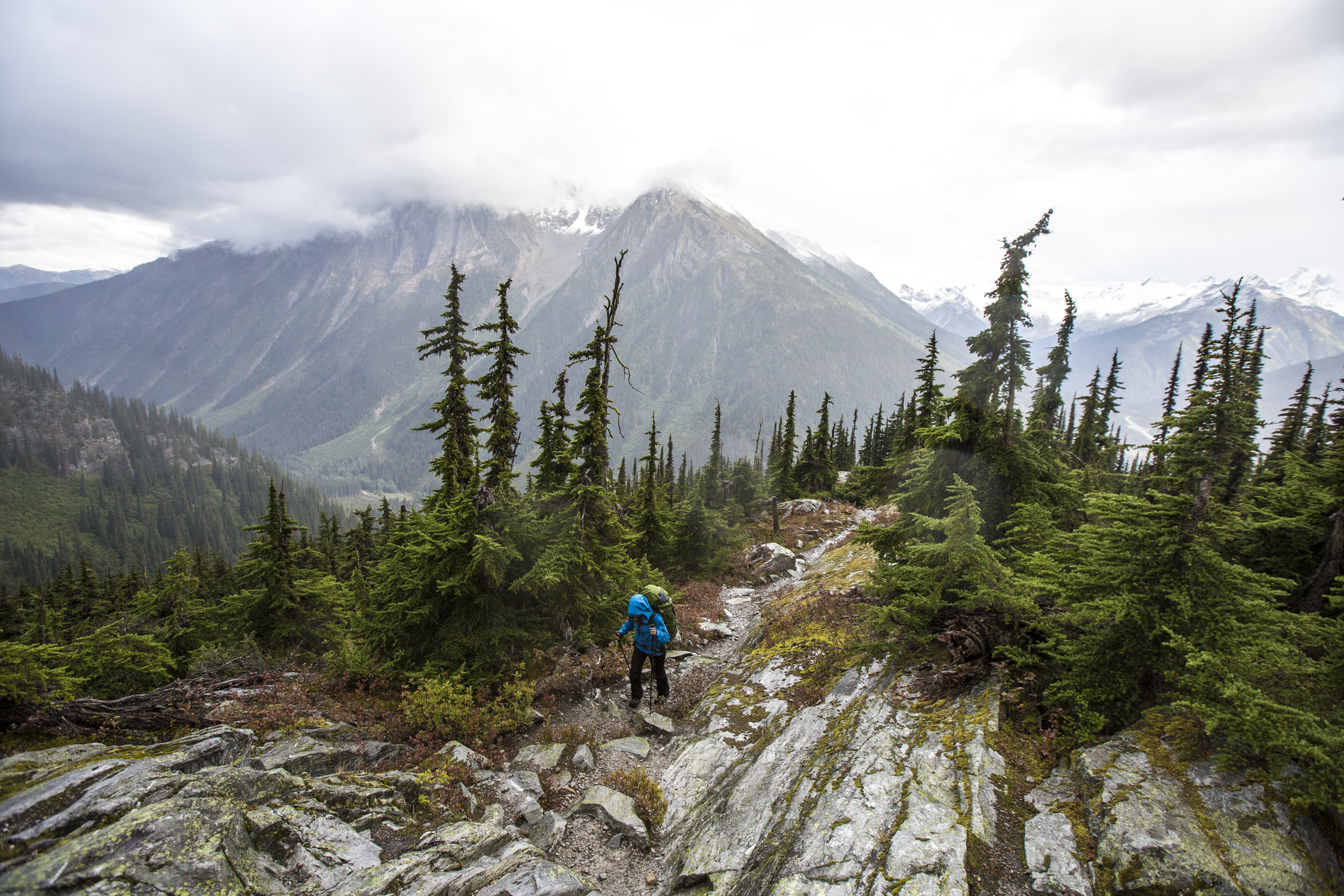 A hiker walks across a rocky outcrop surrounded by trees. They are wearing a blue rain jacket and carrying a green bag. The mountains jut out into the clouds behind them.