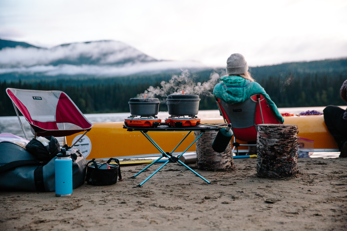 Campers cooking and camping along the banks in Bowron Lake Provincial Park. Two pots are bowling on a camping stove as the campers look out across the lake by their yellow canoe.