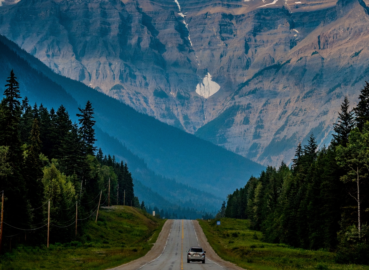 A single car is driving down a road towards Mt. Robson on Highway 16. It's overcast and dark. Evergreen trees line both sides of the highway.