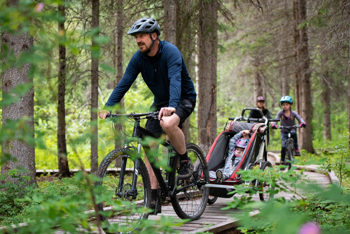 A man pulls a bike trailer with a child inside along a boardwalk bike bath. Two other kids on bikes ride behind him.