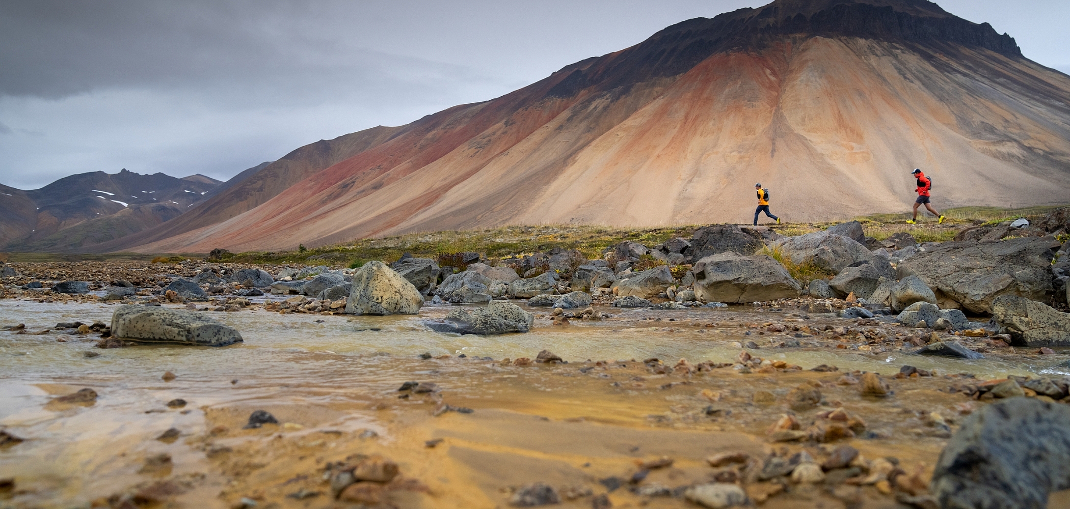 Two trail runners look tiny against the backdrop of a towering Mt. Edziza,streaked with rust and bone colours. 