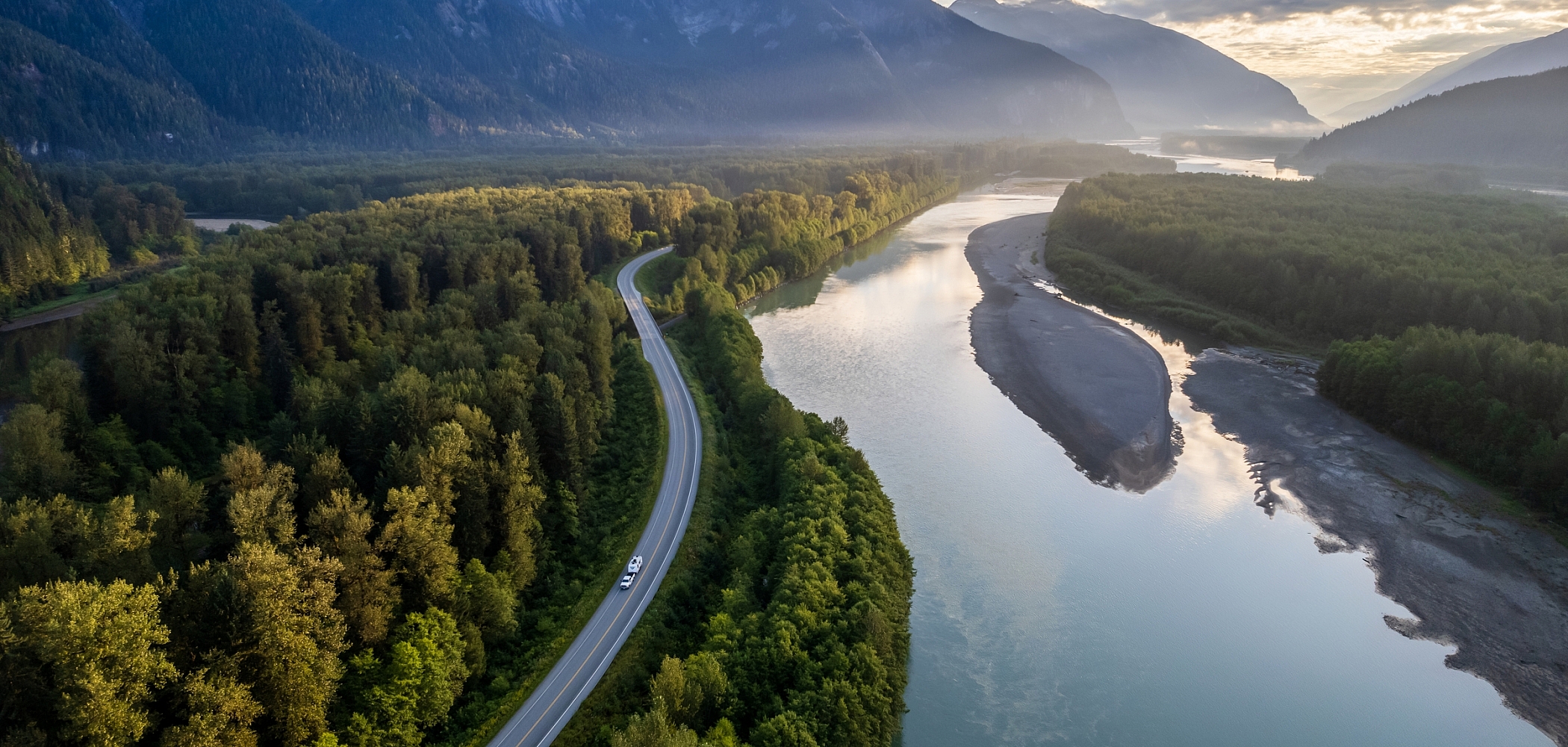 An RV winds its way along a highway, surrounded by dense forest, tall mountains and a calm river. 