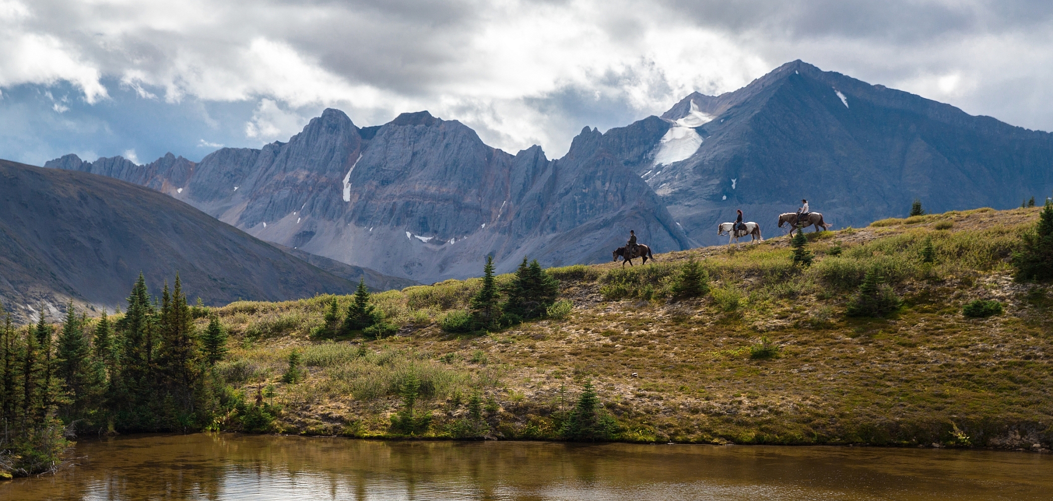 Horseback riders trot along a ridgeline with towering mountains in the backdrop, making them feel tiny in comparison.