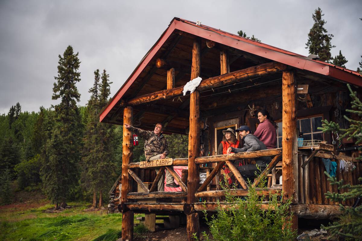 A group of people sits in a small log shelter fishing camp, surrounded by wilderness.